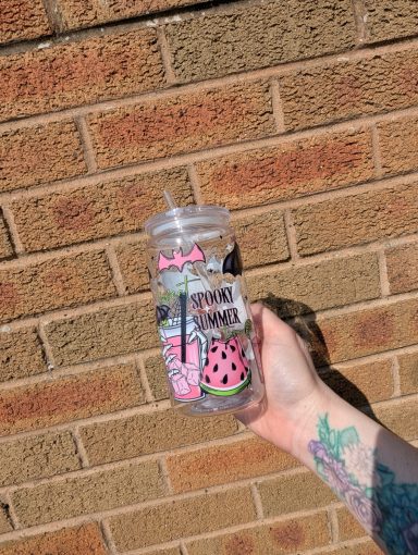 A hand holding a clear cup decorated with watermelons and a pink straw against a brick wall.